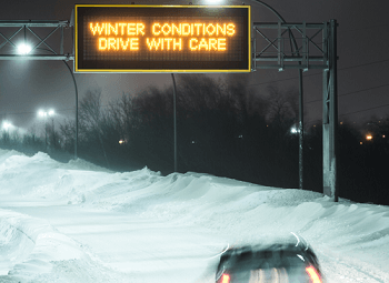 Highway with a lot of snow an overhead electronic sign reads winter conditions drive with care